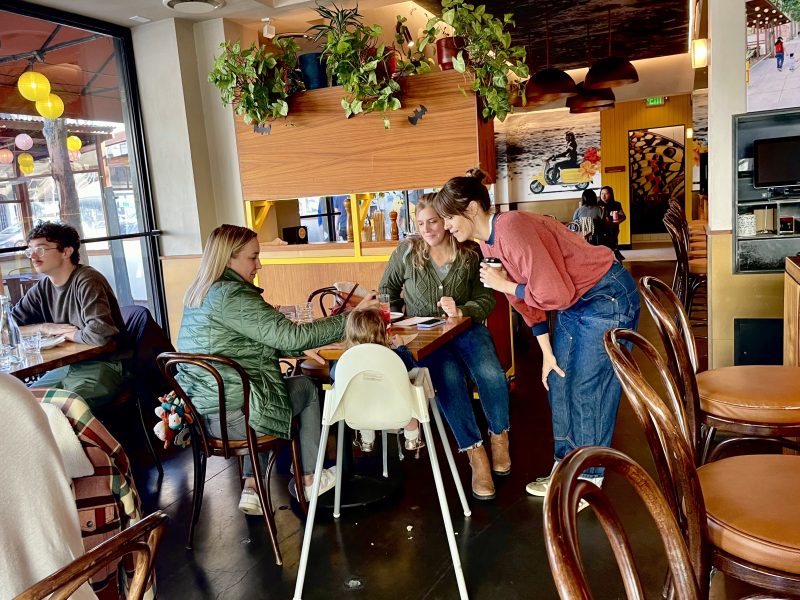 A group of four people, including a child in a high chair, sit at a table in a cozy restaurant with plants hanging overhead.