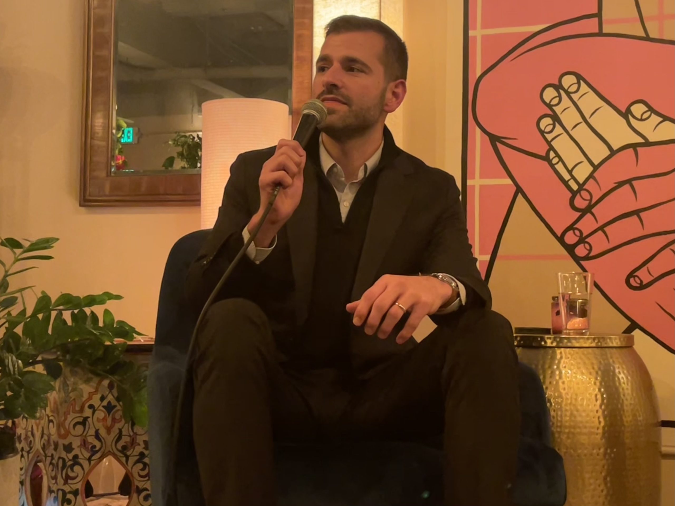A person in a suit sitting on a chair, holding a microphone. There is artwork with clasped hands in the background and a plant next to the chair.