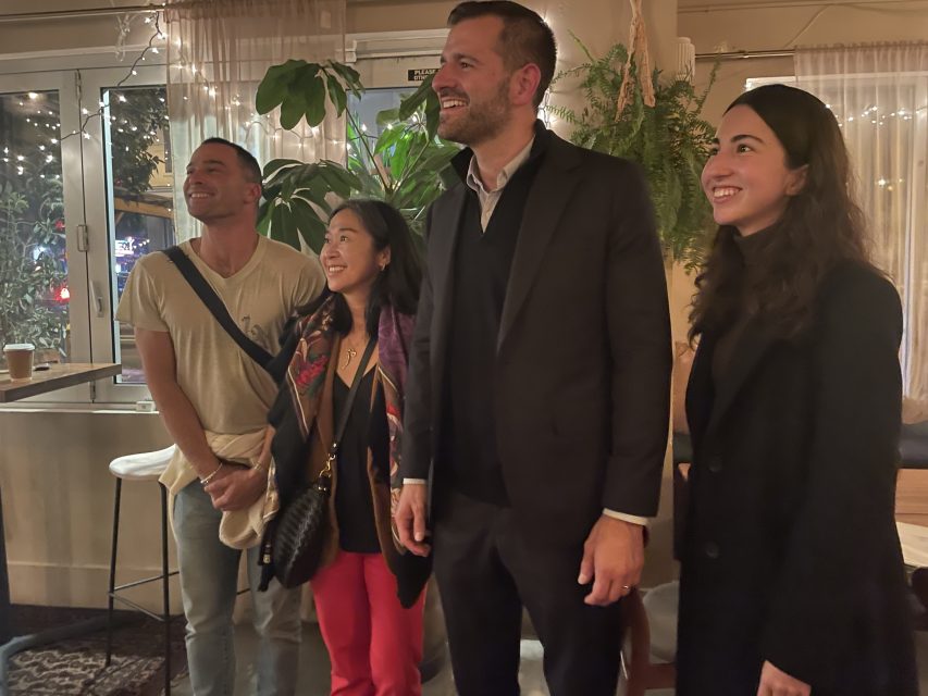 Four people stand smiling in a cozy indoor setting with plants and fairy lights in the background.