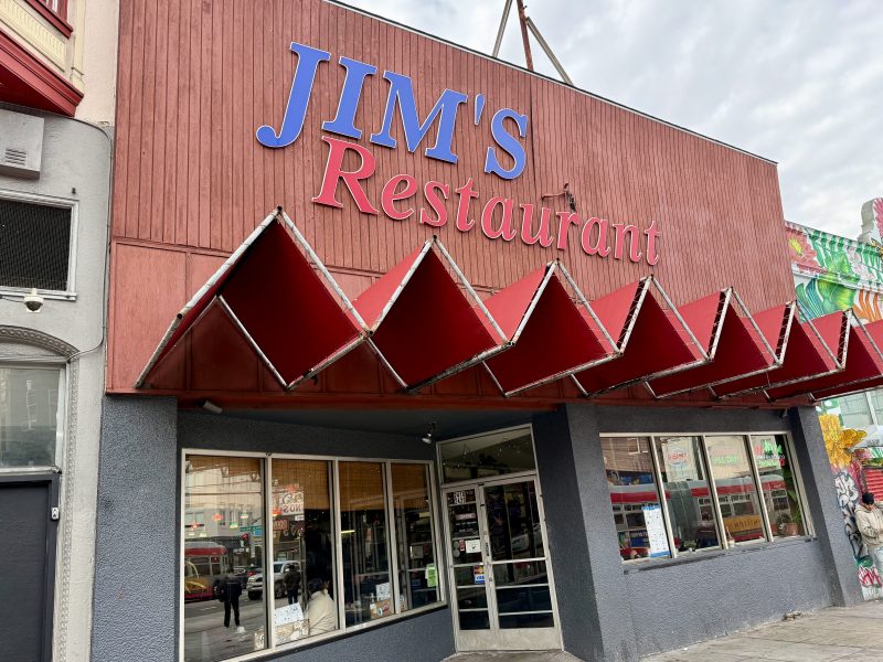 Front view of Jim's Restaurant with a red and blue sign, featuring a geometric awning and street reflections in the windows.