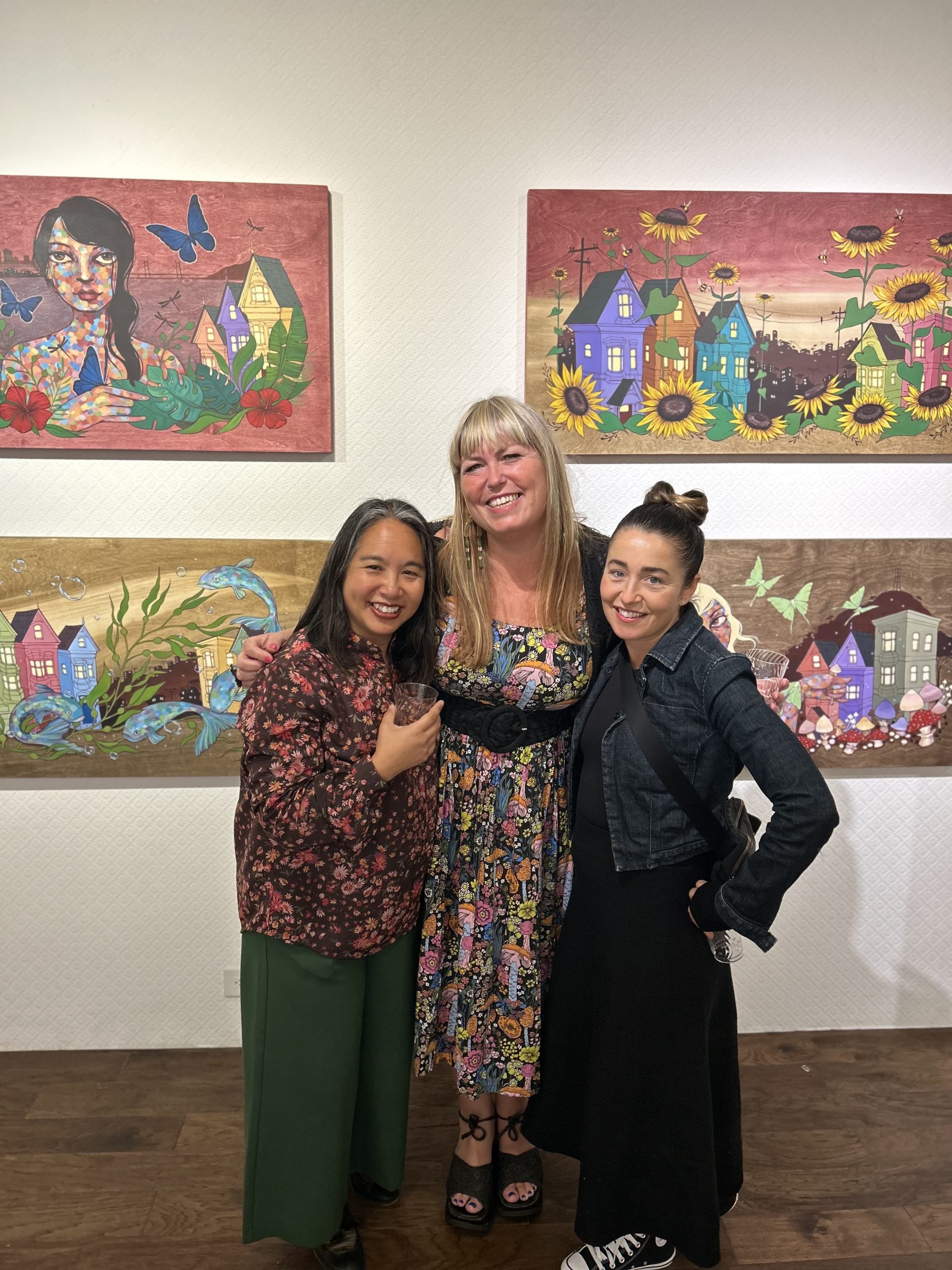 Three people standing in front of colorful paintings in an art gallery, smiling and posing for a photo.