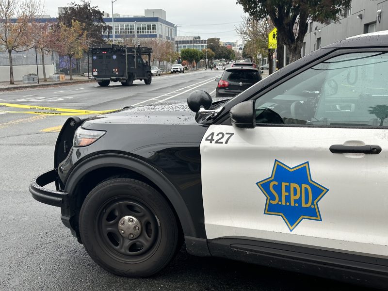 S.F.P.D. vehicles block a rainy street with yellow caution tape. An armored truck is seen in the distance.
