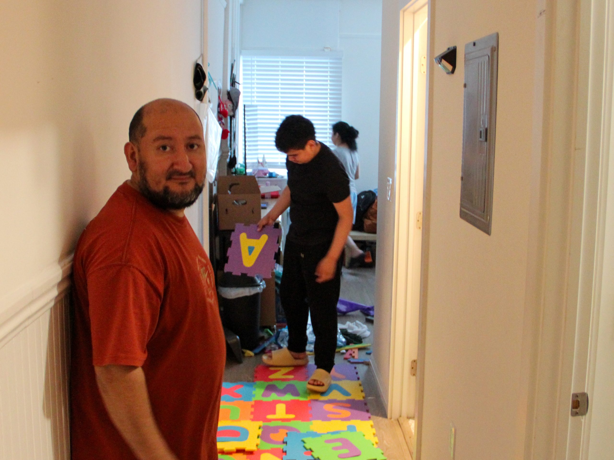 A person in a red shirt stands in a hallway, while another person assembles colorful letter tiles on the floor.