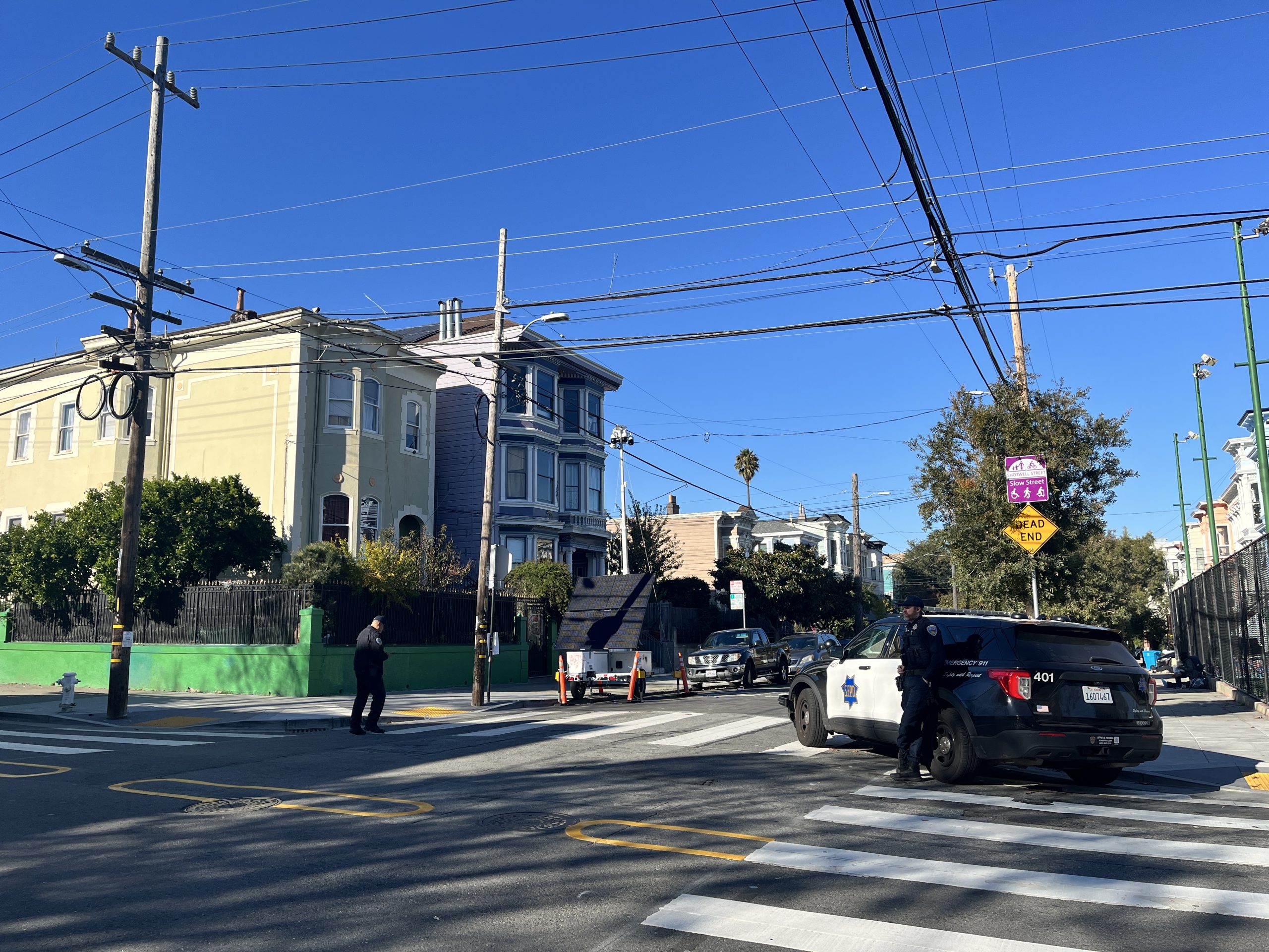 Police vehicles at a crosswalk near residential buildings on a sunny day. Officers are present on the street.