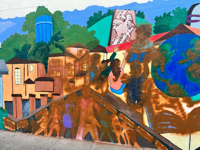 Colorful mural depicting a cityscape with buildings, people, a water tower, and a hand painting a globe.