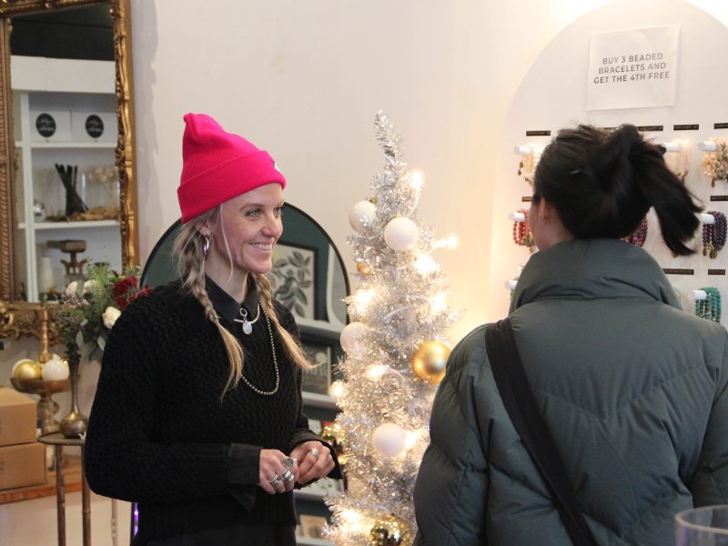 Two people talking in a store with a small decorated Christmas tree in the background. One person wears a pink hat and the other a green jacket. Display of beaded bracelets visible.