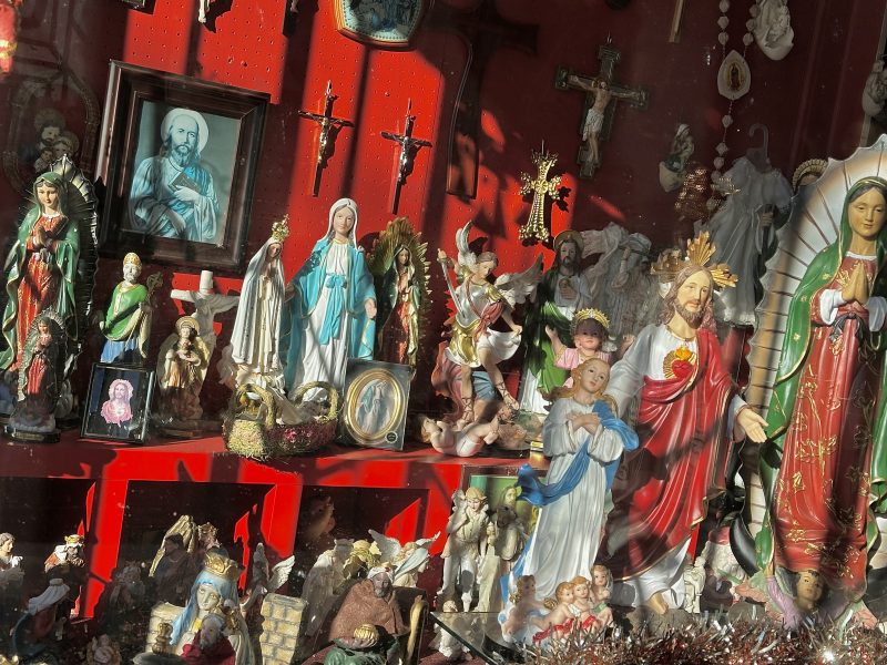 A collection of religious statues and framed images displayed on red shelves, featuring crosses, saints, and depictions of Jesus and Mary.