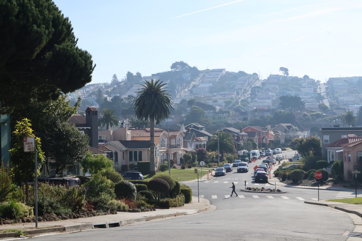 A residential street with parked cars and a pedestrian crossing. Houses line the street, and a hill with more houses is visible in the background.