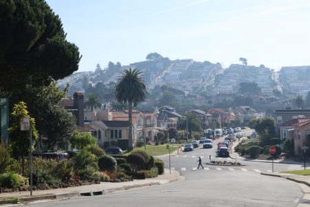 A residential street with parked cars and a pedestrian crossing. Houses line the street, and a hill with more houses is visible in the background.