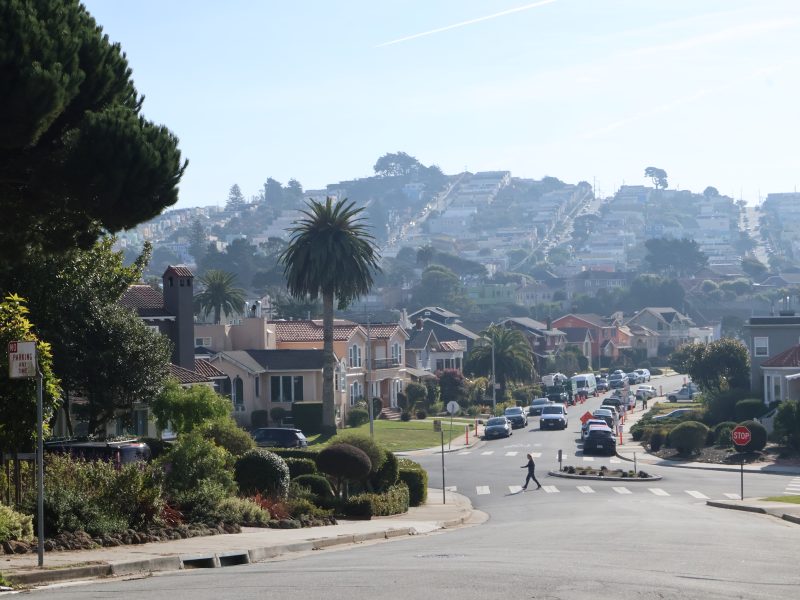 A residential street with parked cars and a pedestrian crossing. Houses line the street, and a hill with more houses is visible in the background.