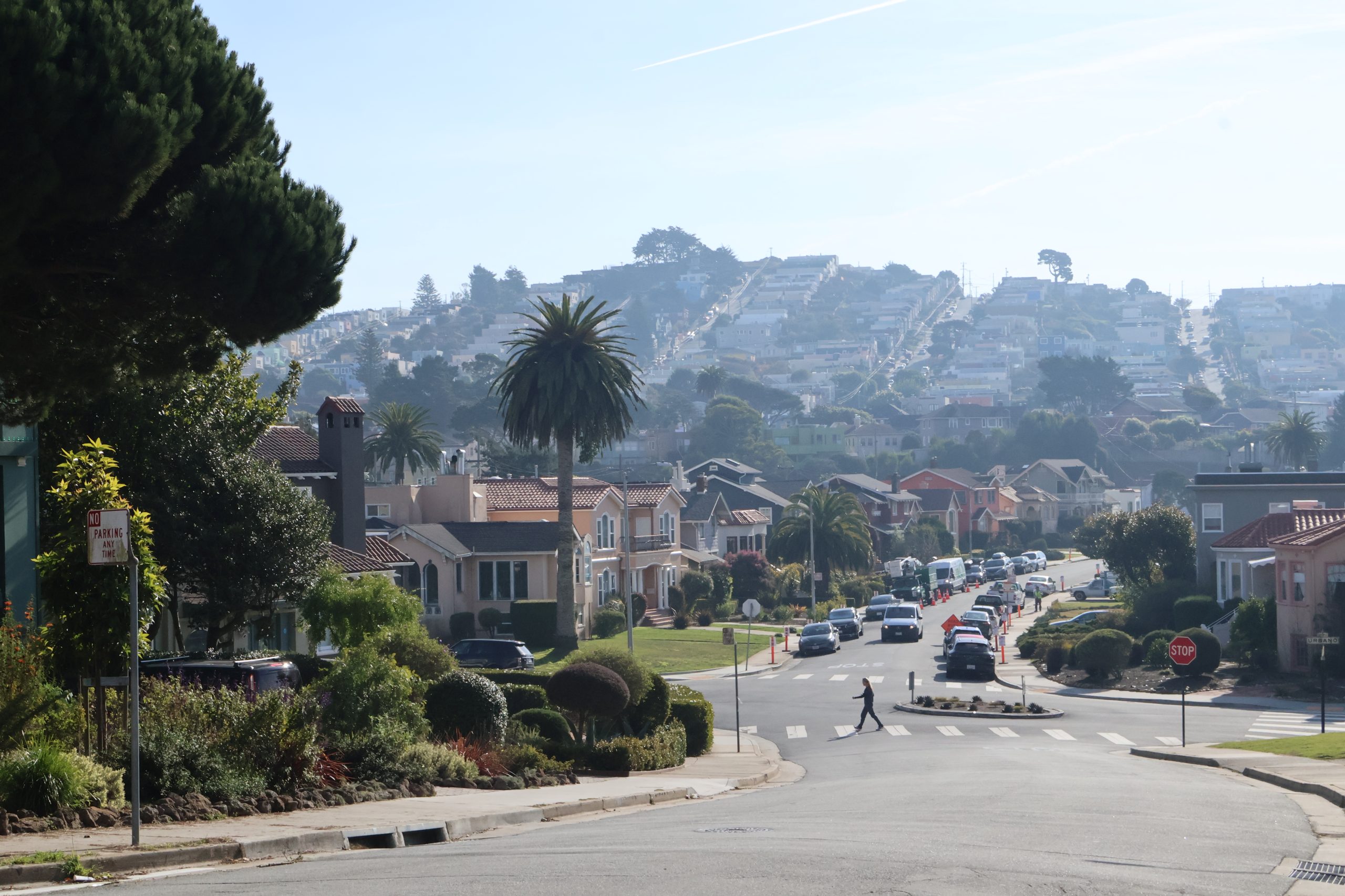 A residential street with parked cars and a pedestrian crossing. Houses line the street, and a hill with more houses is visible in the background.