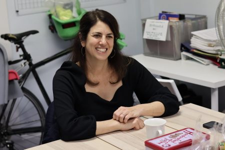 A woman is sitting at a wooden table, smiling. A bicycle is in the background, along with shelves and office supplies. A mug and a red container are on the table.