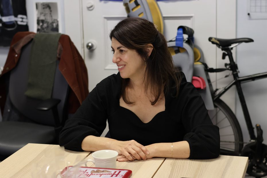 A person with long dark hair sits at a table, smiling and wearing a black top. A bike and chair are visible in the background.