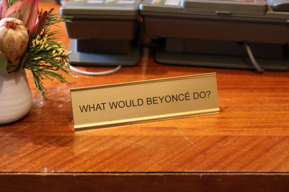 A desk placard reads "WHAT WOULD BEYONCÉ DO?" placed on a wooden surface next to a potted plant and electronic devices.