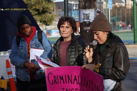 Three people are standing outdoors, one speaking into a microphone. They hold a pink sign with text about criminalization.