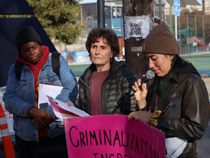 Three people are standing outdoors, one speaking into a microphone. They hold a pink sign with text about criminalization.