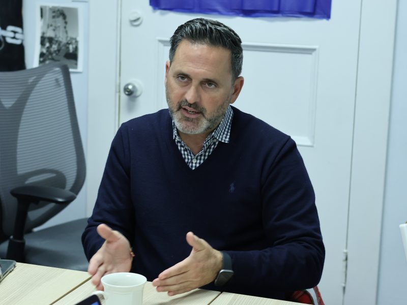 Ahsha Safaí in a navy sweater gestures while sitting at a table with a cup, in an office setting.