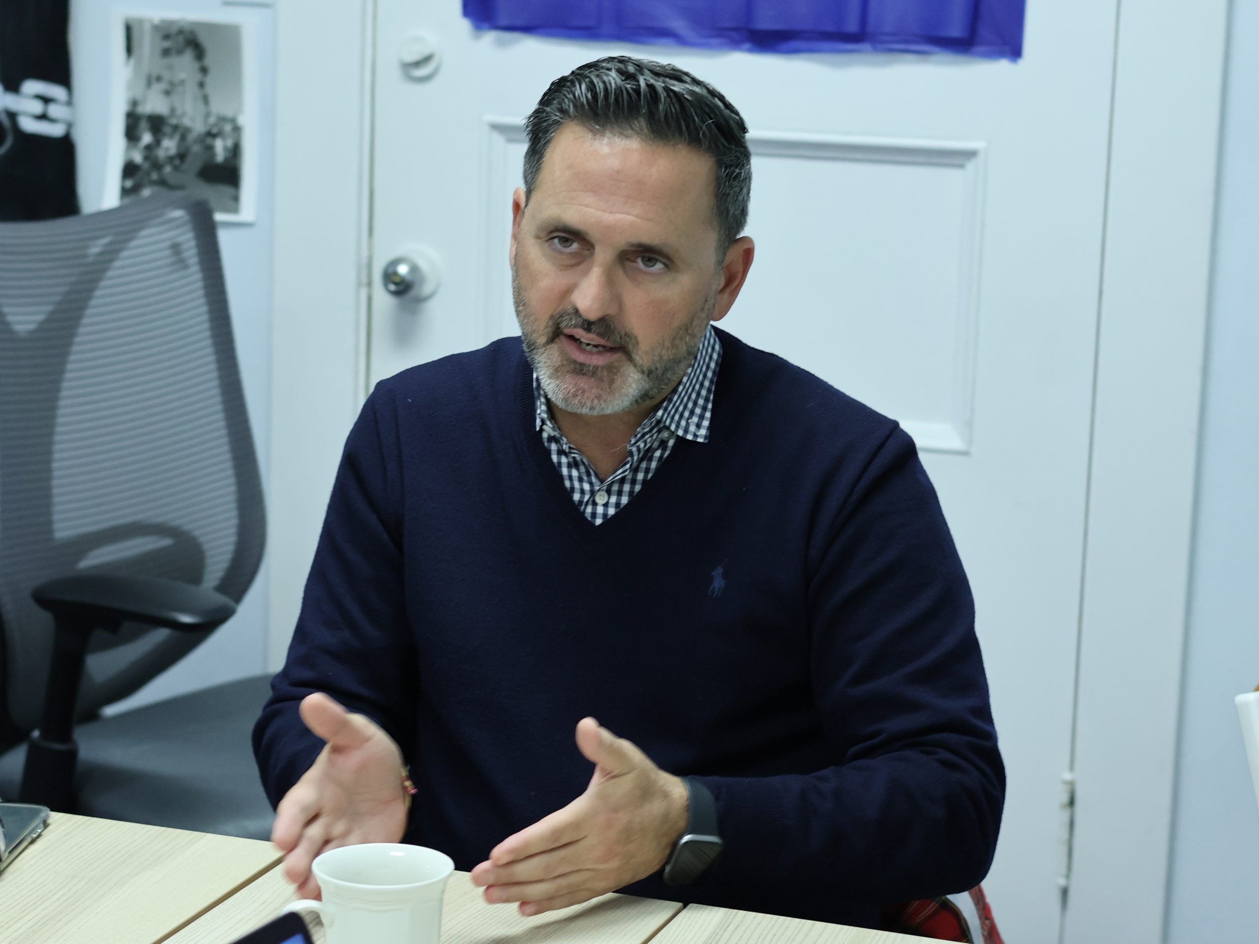 Ahsha Safaí in a navy sweater gestures while sitting at a table with a cup, in an office setting.
