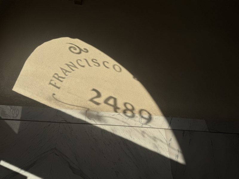 Shadow of the text "FRANCISCO 2480" cast on a wall and tiled floor, illuminated by sunlight.