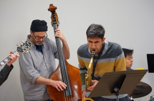 A jazz band performing; one musician plays the double bass, another plays the saxophone, with others playing guitar and piano.