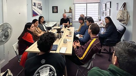 A group of people sit around a conference table engaged in discussion in a small office setting.