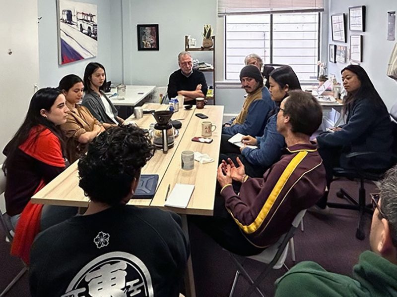 A group of people sit around a conference table engaged in discussion in a small office setting.