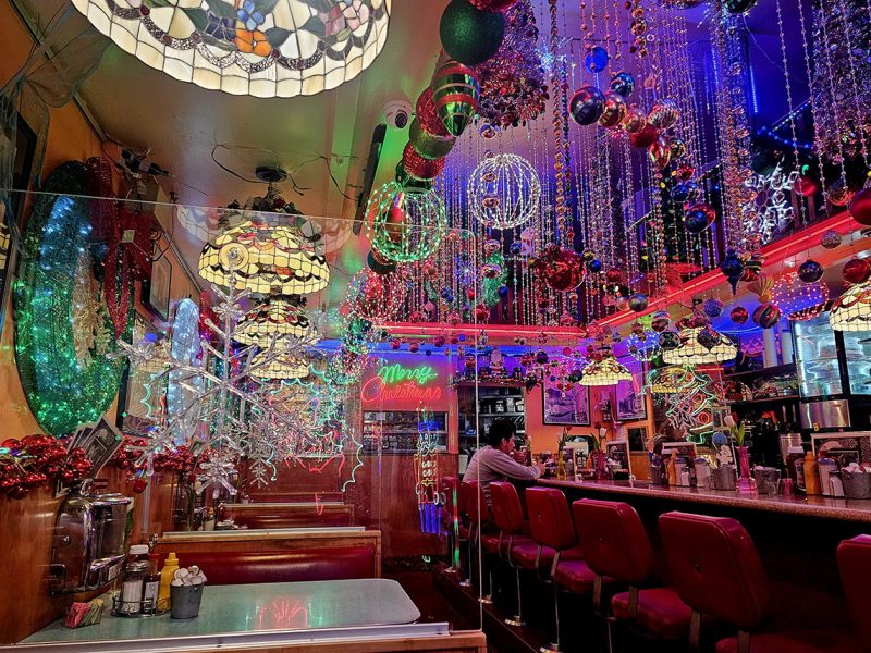 A colorful diner interior decorated with hanging ornaments, Christmas lights, and neon signs. One person sits at the counter surrounded by festive decor.
