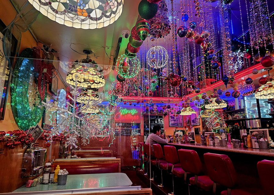 A colorful diner interior decorated with hanging ornaments, Christmas lights, and neon signs. One person sits at the counter surrounded by festive decor.