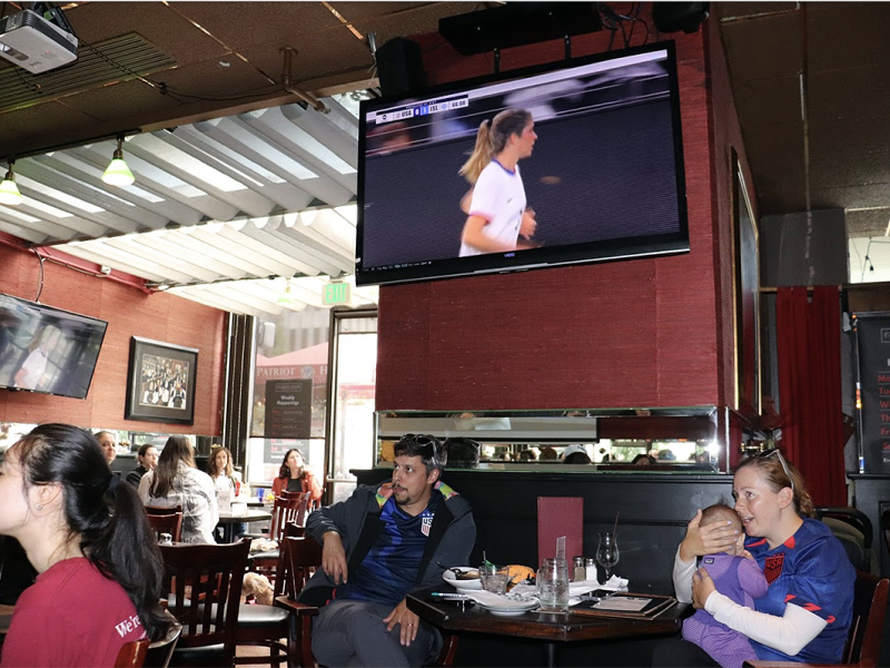 A restaurant with patrons watching a sports game on two TVs. A woman holds a baby at a table while others sit and chat.