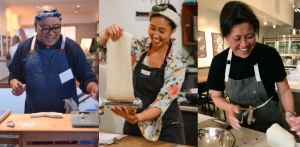 Three people making pasta: one is kneading dough, another is using a pasta machine, and the third is smiling with hands in dough. They are in a kitchen setting, each wearing an apron.