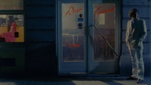 A person in a white suit stands outside a dimly lit restaurant with "Rose" and "Greneral" written on the glass door. Another person is seated inside.
