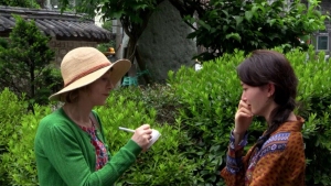 Two people are having a conversation outdoors near green foliage. One person is holding a notepad and pen, and the other is touching their face thoughtfully.