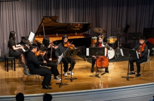 A small chamber ensemble performs on stage with a grand piano, violinists, a cellist, and a drummer.