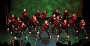 A dance group performs energetically on stage, wearing red and black outfits, under green lighting.