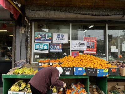 A person selects produce from a market stand displaying bananas, oranges, and avocados. Campaign posters are visible on the window above.