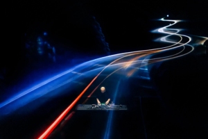 A person stands at a DJ console under a dark sky with colorful light trails curving above.