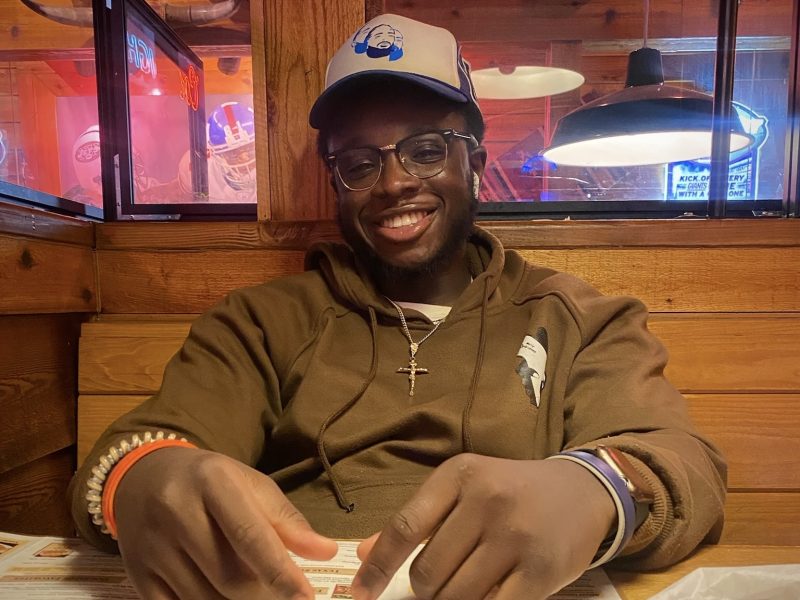 A person in a brown hoodie and cap sits at a wooden table, smiling and holding a menu in a warmly lit restaurant.
