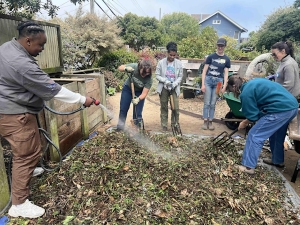 A group of five people is gardening, watering, and tending to a large compost bin outdoors.