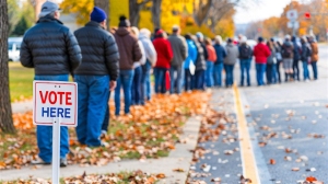A long line of people waits to vote, with a "Vote Here" sign in the foreground and autumn leaves scattered on the ground.