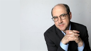 A person with glasses and a suit is seated against a plain background, looking at the camera with hands clasped.