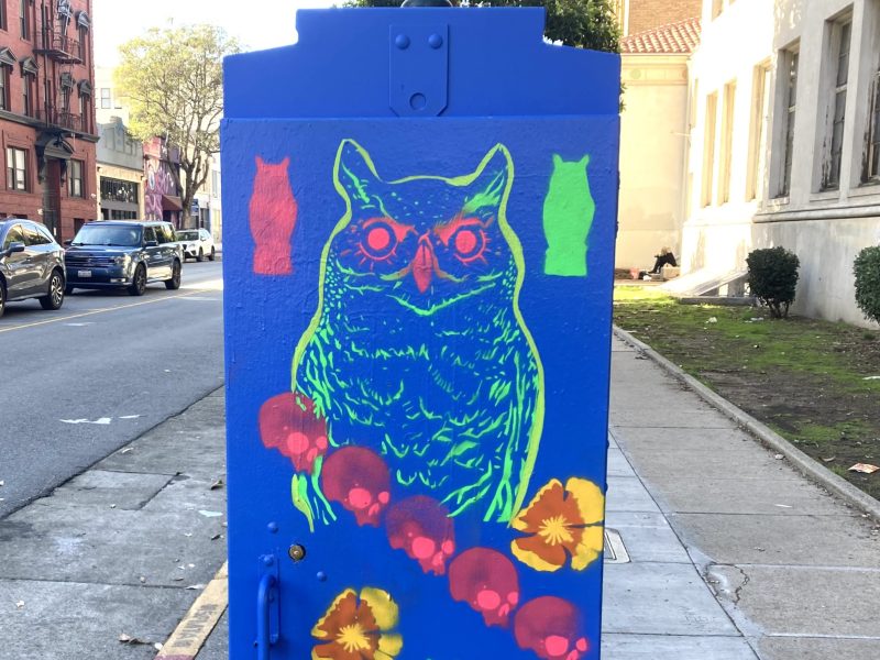 Bright street art features a neon green owl with red eyes on a blue background, surrounded by colorful flowers and ghostly shapes, painted on a utility box by the sidewalk.