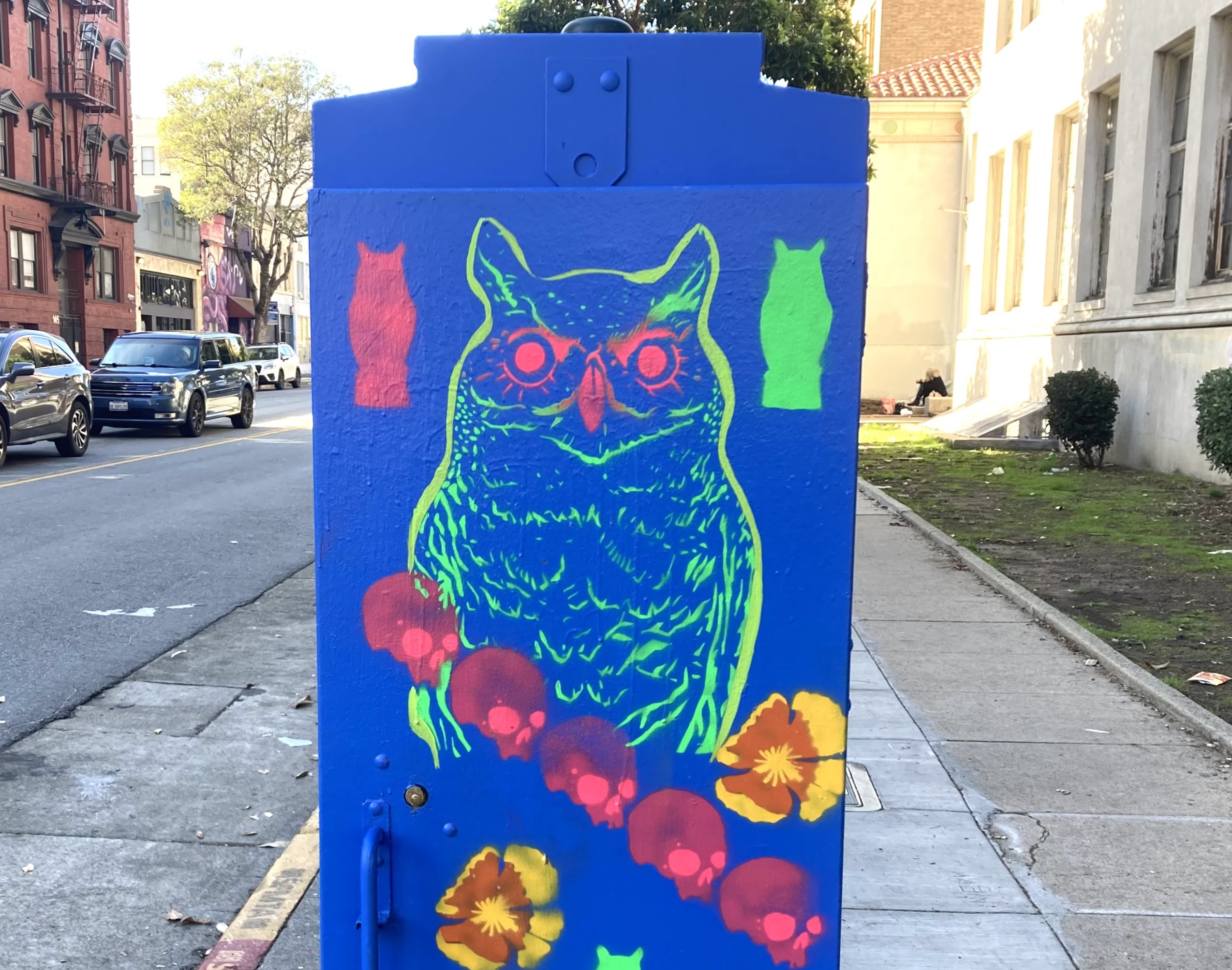 Bright street art features a neon green owl with red eyes on a blue background, surrounded by colorful flowers and ghostly shapes, painted on a utility box by the sidewalk.