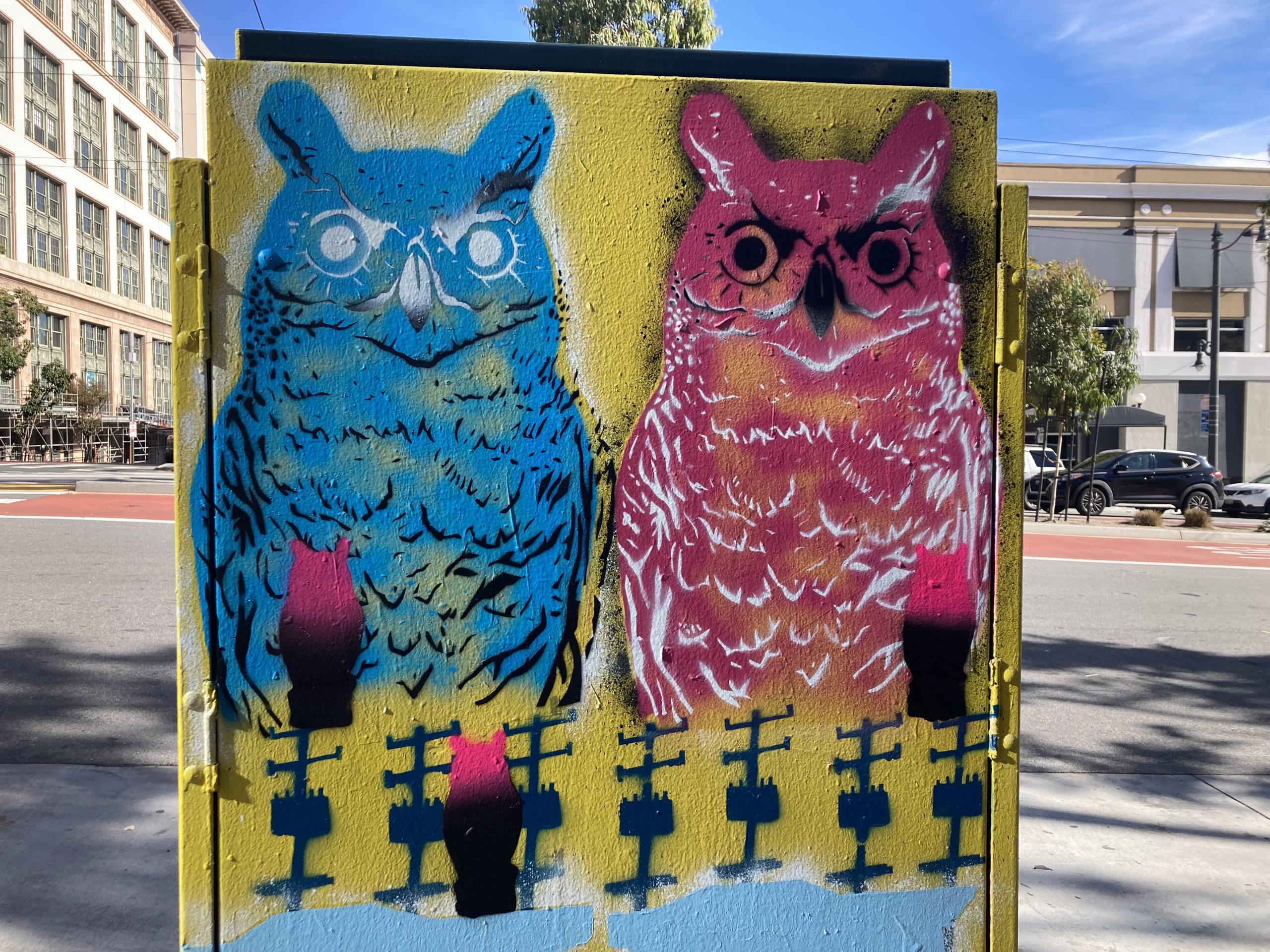 Colorful graffiti of a blue and a pink owl on a utility box, with a yellow background and black fence design at the bottom.