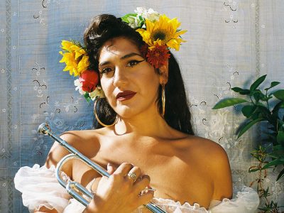 Person with flowers in their hair holds a musical instrument, wearing large hoop earrings and an off-the-shoulder top, with a neutral expression.