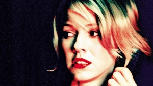 A person with short blond hair and red lipstick looks to the side against a dark background.