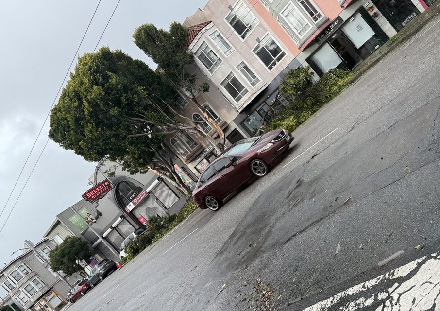 A maroon car is parked on a city street, framed by buildings and a tree lining the sidewalk. The overcast sky looms above, hinting at an impending storm.