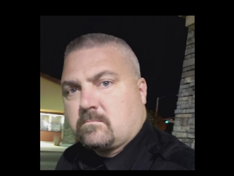 A man with short hair and a goatee is standing outdoors at night, looking at the camera. There's a building and a stone pillar in the background.