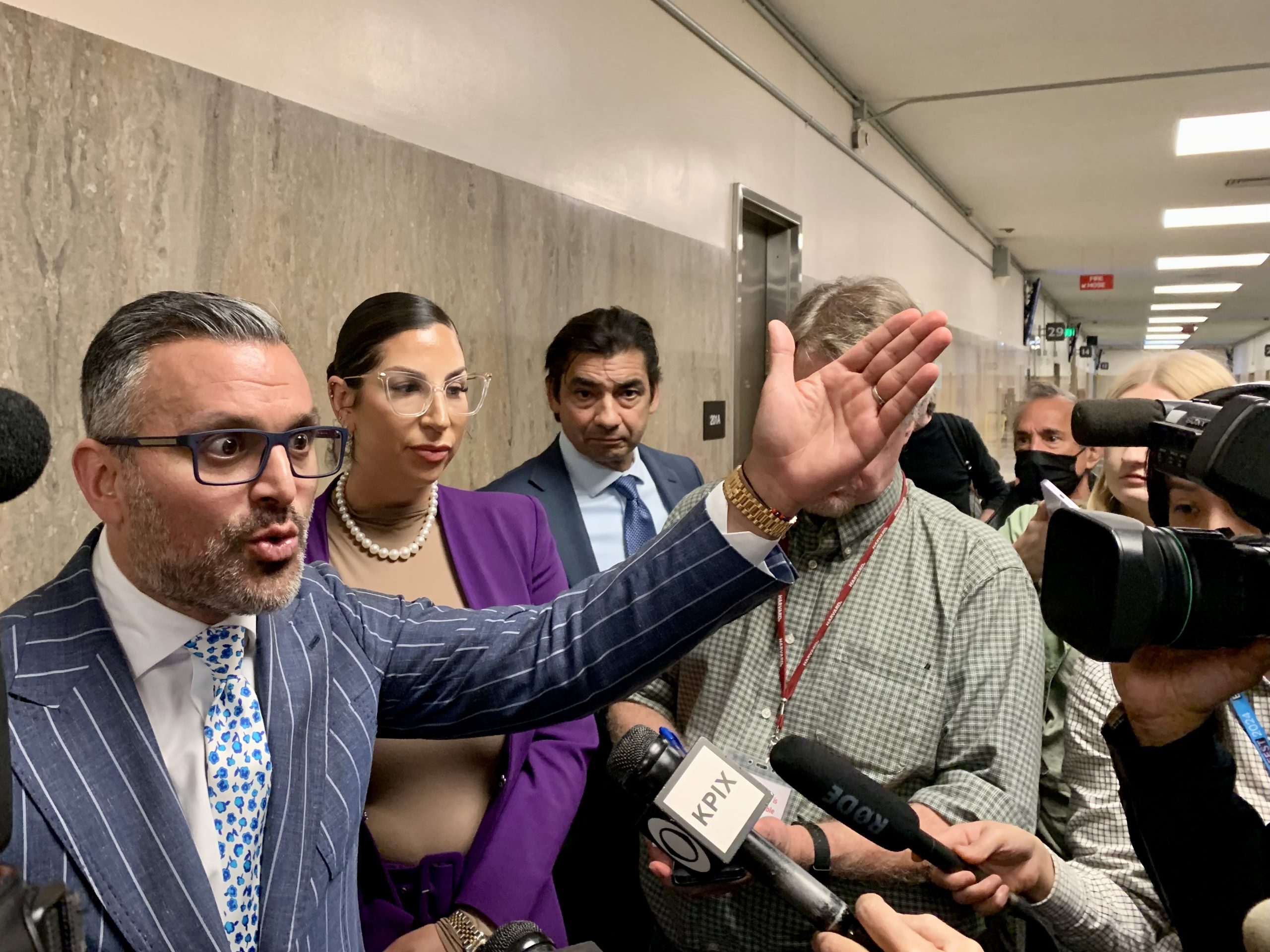 A man gestures while speaking to reporters in a hallway. Microphones are directed at him, and several people stand nearby, including a woman in glasses and a man in a suit.