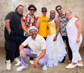A group of seven people pose in colorful and casual clothing against a textured wall.