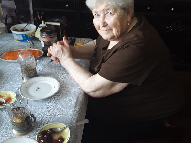 Elderly woman sitting at a dining table with various dishes and jars, looking at the camera.